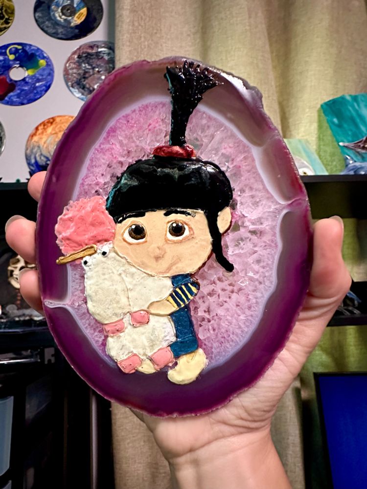 A glaze painting on polished pink agate depicting Agnes from Despicable Me hugging a fluffy plush unicorn. 