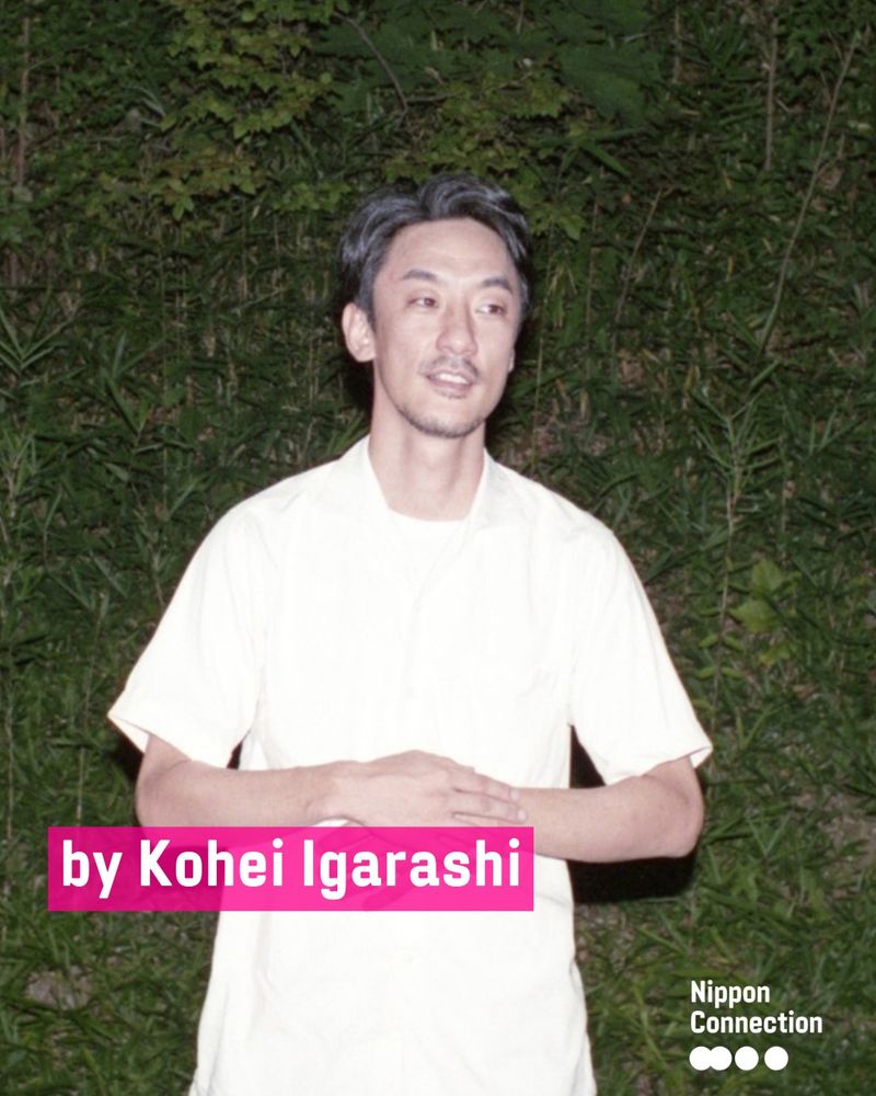 Porträt des Regisseurs Kohei Igarashi: Er steht in einem weißen Hemd vor grünem Blätterhintergrund und blickt leicht lächelnd zur Seite.