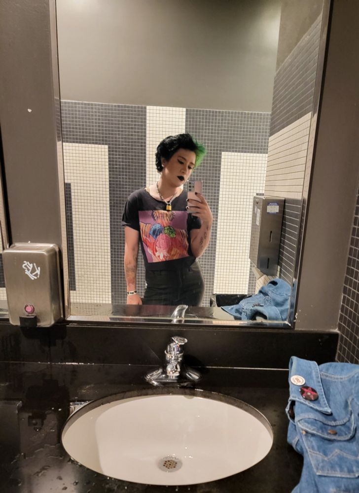 A guy with green and black hair poses for a selfie in a bathroom