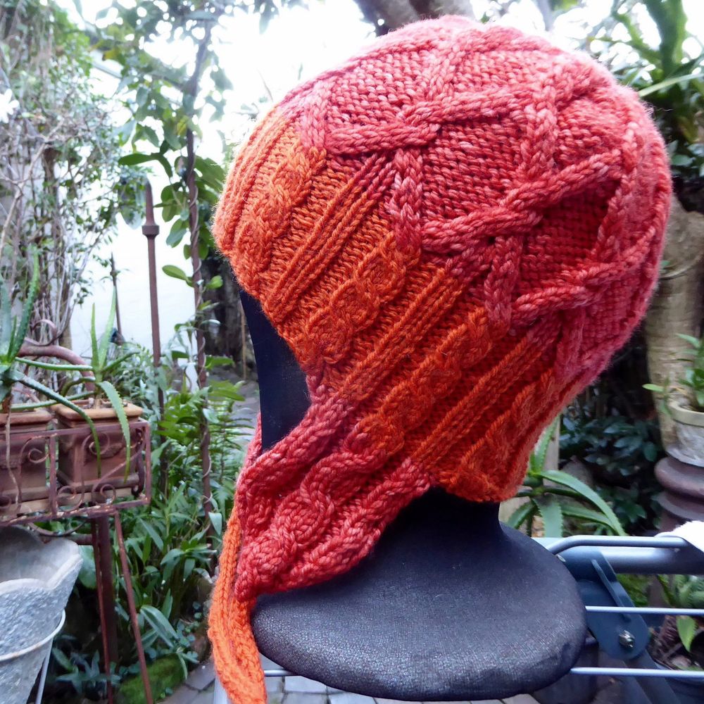 This photo taken in my garden so various plants and pots etc are in the background.  

A bright orange & red orange beanie hat with ear-flaps is shown on a styrofoam ‘head’ that’s been covered with a black stocking. 

The hat has Lattice design cabling on the head - the wide band is ribbing but every 2nd knitwise rib has been replaced with a thin twisted rope cable … that extends into the ear flaps which then taper into the ties. 

I made this for a friend - adapted from the pattern ‘Merry’ by Amanda Schwabe on Ravelry. Yarn was by Manos del Uruguay (their Gloria - it’s now discontinued) and Bendigo Woollen Mills 8ply