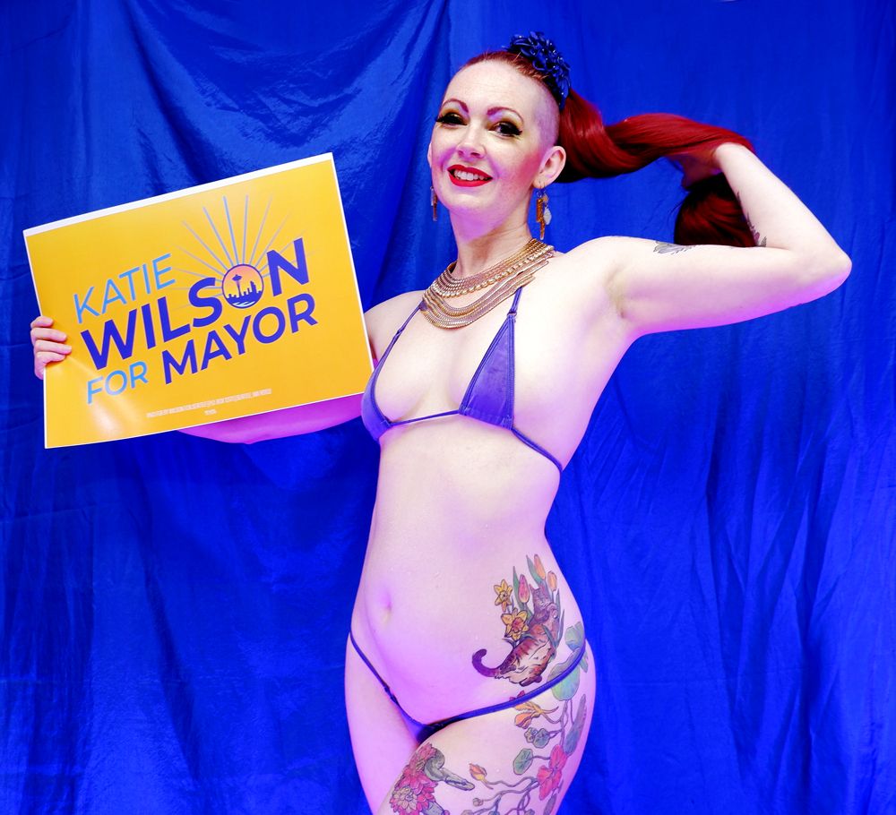A pinup of a pale white woman in a very small blue string bikini and a long red ponytail posing with a Katie Wilson for Mayor sign.