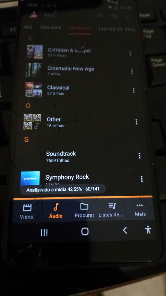 Foto de telefone Galaxy A01 Core, tela rachada, faltando pedacinhos da tela. O VLC está aberto, na ala de Áudio seção Gêneros. As opções a mostra: Christian & Gospel - 537 trilhas (de enfeite); Cinematic New Age - 1 trilha (a Samsung faz bons jingles); Clasical - 67 trilhas (de Clássica não tem muita nesse Classical); Other - 16 trilhas (Michael Jackson tá ali pra não criar outra opção); Soundtrack - 7659 trilhas (e aumentando); Symphony Rock - 1 trilha (a Samsung faz bons jingles). Abaixo na tela há uma barra laranja e uma mensagem "Analisando a mídia 42,55% • 60/141".