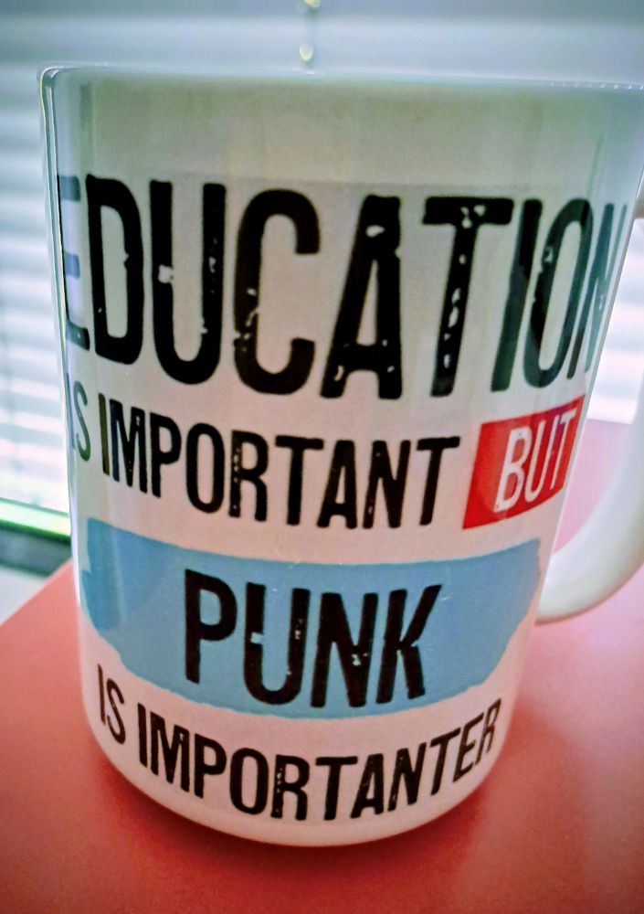 Eine weiße große Tasse auf der steht education ist important but punk is importanter
Die Schrift ist schwarz alles ist in Groß und Buchstaben geschrieben das Wort but ist rot unterlegt das Wort Punk ist hellblau unterlegt

Die Tasse steht auf einem flamingofarbenen untergrund im Hintergrund sieht man verschwommen geschlossene Lamellen an einem Fenster