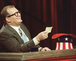 comedian drew carey as the host of the american version of "who's line is it anyway?".  He's wearing a dark gray suit, seated behind a desk, and is smiling in the direction of the performers as he reads a prompt written on a yellow post-it note, pulled out of a hat embellished with the american flag 
