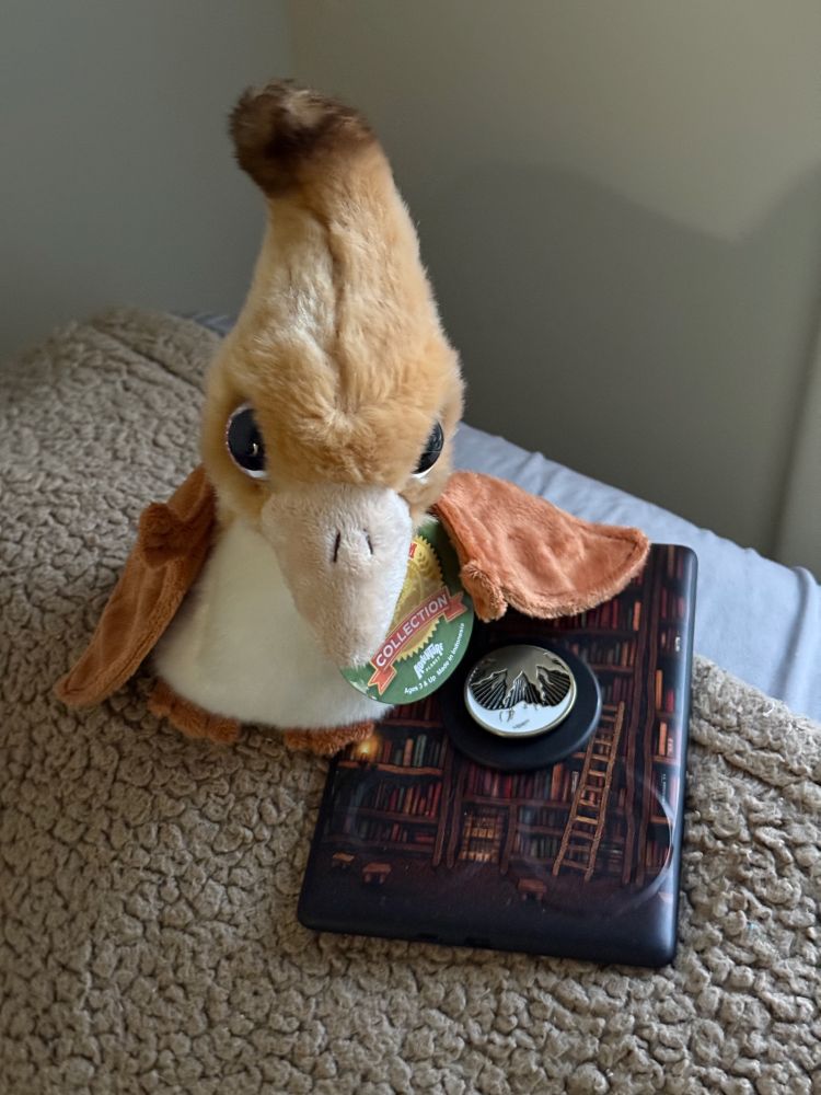 a brown plush pteranodon, leaning on a kindle reader, sitting on a brown blanket