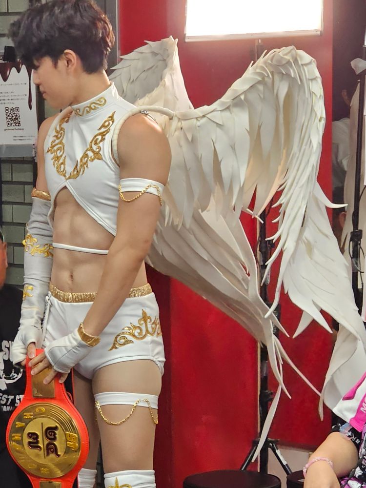 Monomoth in all white with big angel wings and intimidatingly defined abs, demure standing by the door of Ichigaya Chocolate Square holding the SetUp 24/7 Championship.