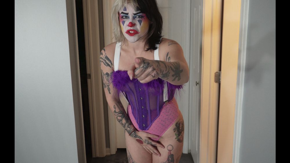 Devious Skunk is wearing a purple fluffy corset with white suspenders attached to pink polka dot underwear. She's wearing clown makeup and covers her panties as she angrily points at the camera