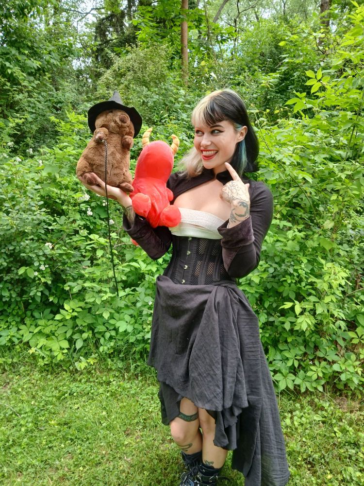 Devious Skunk wearing a black skirt, bustier, and boots holding a stuffed orange dragon and a stuffed Quokka wearing a wizard hat and holding a tiny whip.