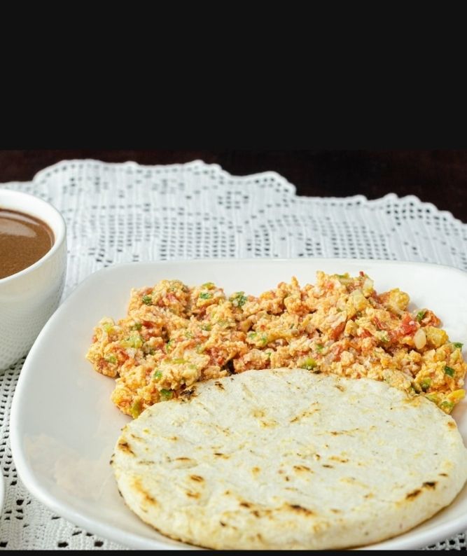 Arepa sobre plato blanco, al lado tiene huevos revueltos con un guiso de tomate, cebolla y perejil
