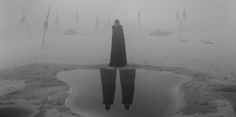 Image is a black and white still from Joel Coen's 2021 film, The Tragedy of Macbeth. It shows a cloaked figure standing at the edge of a pond, looking down at a reflection of two robed figures looking back at them. In the background, mist obscures what looks like a battlefield with torn banners and bodies littering the landscape.