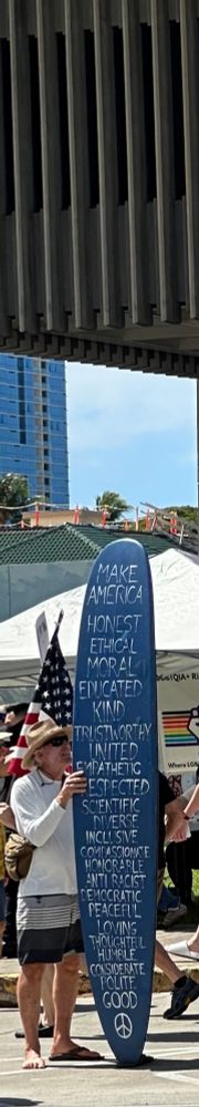A man holding a surfboard that’s painted blue. The words are painted in white & one atop the other. The words are: Make America Honest, Ethical, Moral, Educated, Kind, Trustworthy, United, Empathetic, Respected, Scientific, Diverse, Inclusive, Compassionate, Honorable, Anti Racist, Democratic, Peaceful, Loving, Thoughtful, Humble, Considerate, Polite, Good. At the very bottom is a peace sign ☮️ 