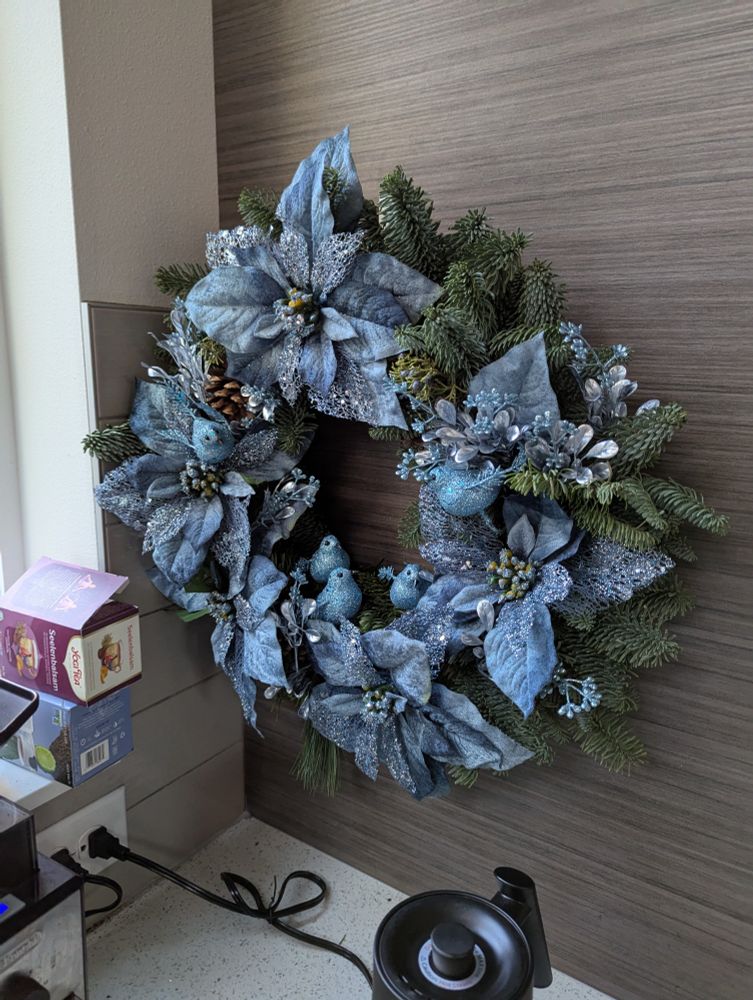 A festive wreath, adorned with blue flowers, with blue glittery bird models camouflaged amongst the other colorful foliage. 