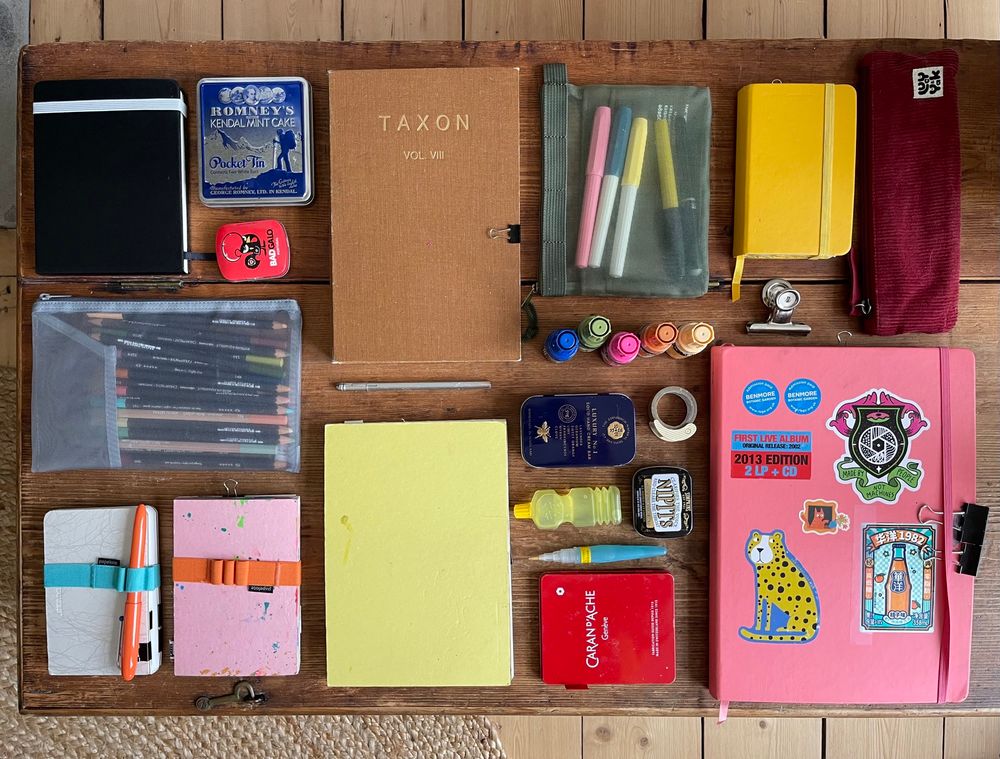 A flatlay of way too many sketchbooks of various sizes and tins and pencil cases filled with paints, ink, crayons, pens and pencils