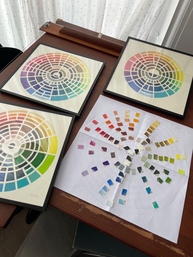 A draftsman table with three framed color wheels and one in the making: small paper samples of colors spread on a glued together large color wheels schematics. Starting at 12 colors go from yellow to green to teals to blues at 6 to purples to reds to skin tones and oranges. Blacks and whites are in the center. Colors go from dark in the middle to light at the edges.