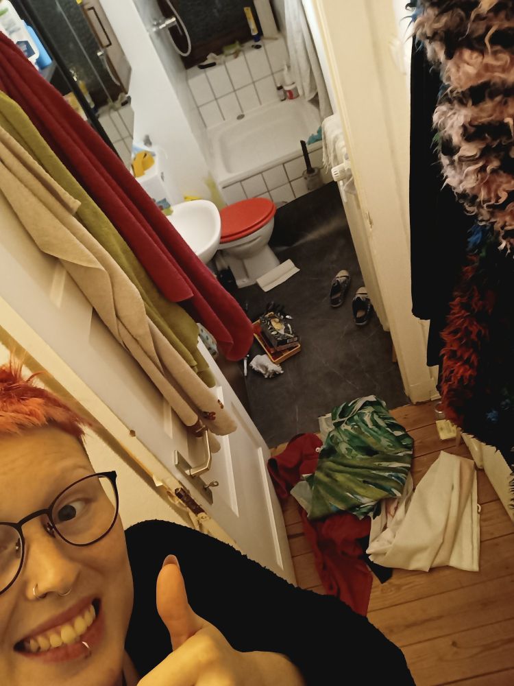 unten links grinst eine person in die kamera und hält den daumen hoch. im Hintergrund sieht man ein chaotisches kleines badezimmer mit werkzeug und handtüchern auf dem boden verteilt.