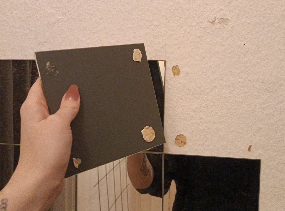 hand hält abgelöstes Spiegelstück mit resten der tapete an der Rückseite. daneben ist die wand mit fehlender tapete