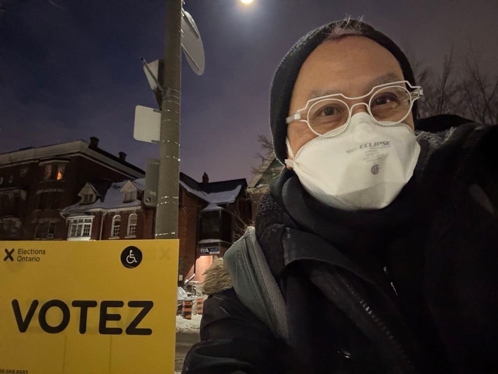 selfie of me wearing white glasses, a white n95 mask, black toque and black winter coat, standing in front of a yellow elections ontario VOTEZ sign at night 