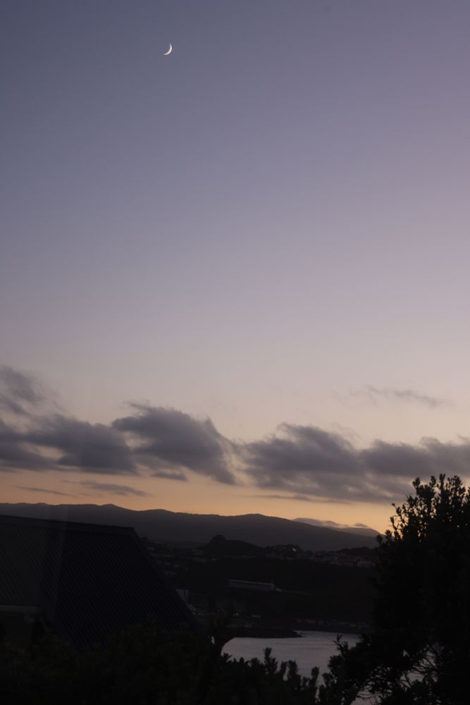 A crescent moon at dusk above low clouds and distant hills in Te Whanganui a Tara.
