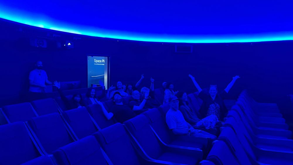Inside the planetarium, the Te Wānanga o Hina group smiling and waving on the seats beneath the dome, lit by blue light.