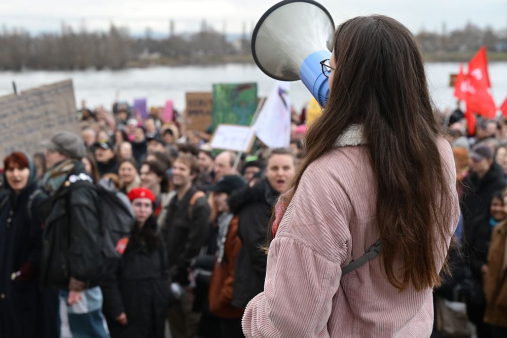 Eine Person mit Megafon ruft Parolen. Im Hintergrund sind Kundgebungsteilnehmende zu sehen.