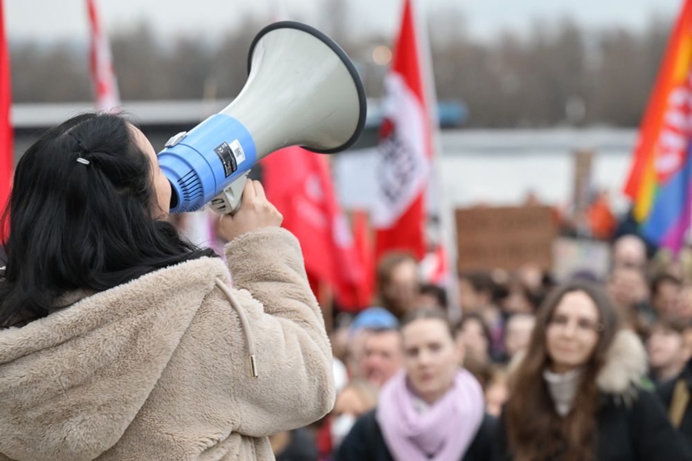 Eine Person mit Megafon im Vordergrund. Im Hintergrund sind Teilnehmende der Kundgebung zu sehen.