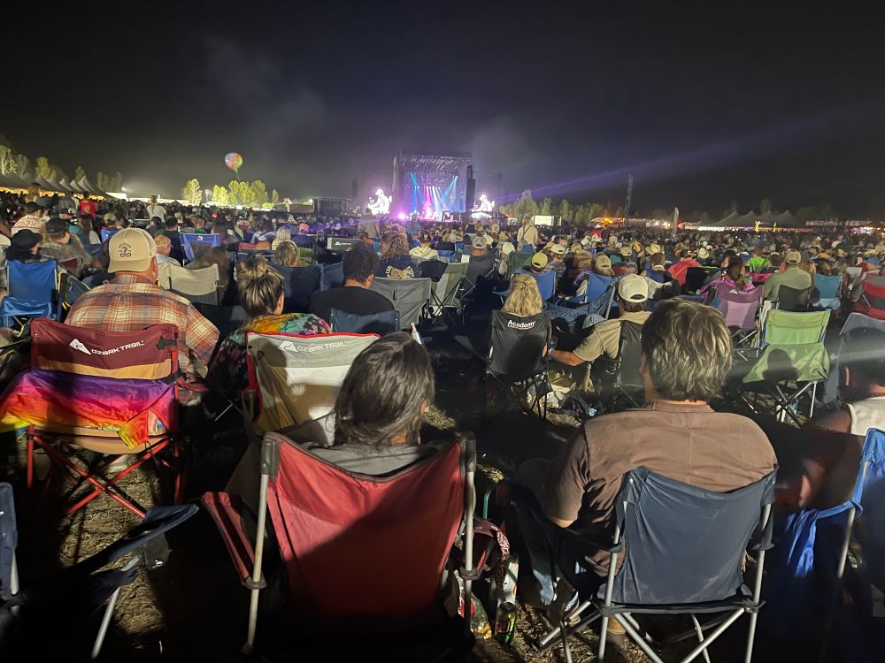 Night scene with a large crowd of people in camping chairs and a brightly lit stage in the distance