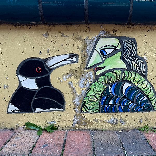 Street mural of a magpie speaking with an old lady with green skin who may or may not be a witch (she has a pointy nose)