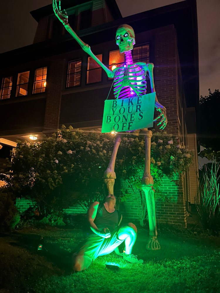 A giant skeleton with a sign saying “bike your bones off” im crouching in front miming jorking it