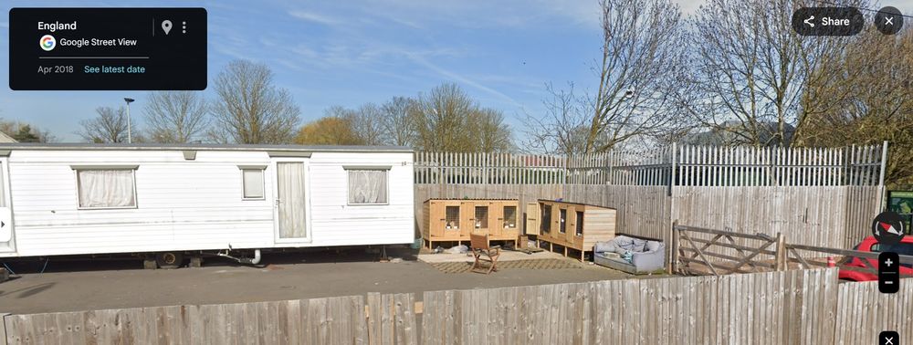 A caravan sits next to two three-cage wooden chicken boxes - a chair is set up facing them, and a couch next to one of them, in a concrete yard. 