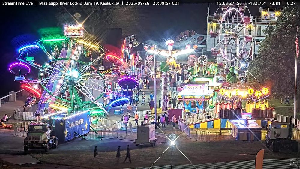 At 8 p.m. on September 26th the figures of a few people in the foreground appear tiny under the blaze of colorful neon lights on more than seven rides filling the middle ground, and the glow of the waxing moon highlights the river flowing by in the background. 