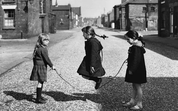 Two young girls standing in the road hold a skipping rope, a third girl jumps over the rope, her ponytail flying up.