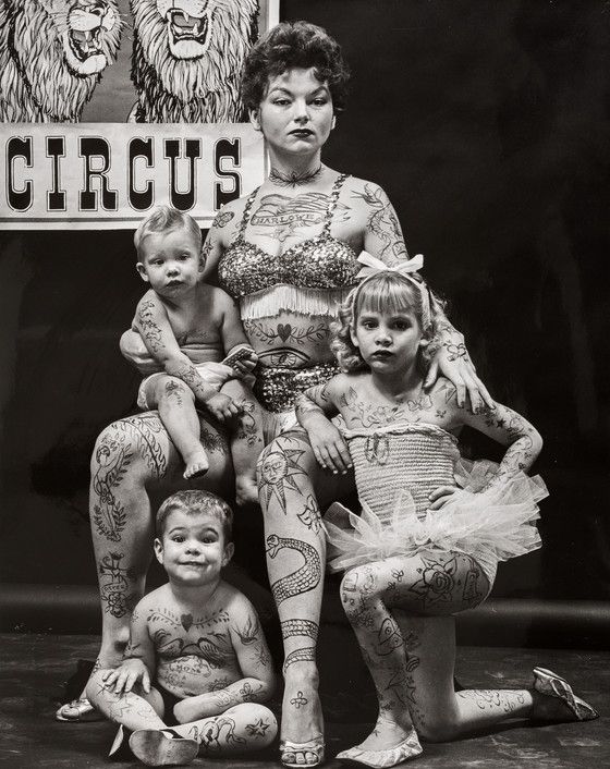 A family of American circus performers. A mother and her three young children. All appear to be heavily tattooed. The mother looks proud and no-nonsense, an expression her blonde daughter also shares. The boy sitting cross-legged on the floor is smiling and the baby on his mom's knee looks somewhat puzzled. Behind them is a poster that reads Circus and shows two cartoon lions.