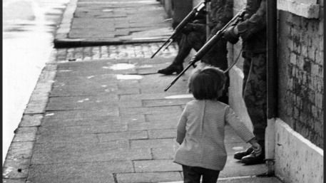 a tiny child walks away down the street towards the guns of two British soldiers sticking downwards out of the doorways in the street.