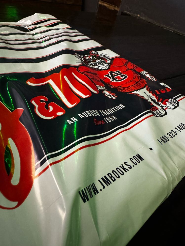 The image shows a plastic bag from “J&M,” an Auburn University tradition since 1953. The bag features an illustration of a tiger mascot wearing an “AU” sweater, standing in a spirited pose. The text on the bag includes the store’s website, “www.jmbooks.com,” and a phone number, “1-800-323-1409.” The bag has a vintage design style with black, white, and red stripes across the top.