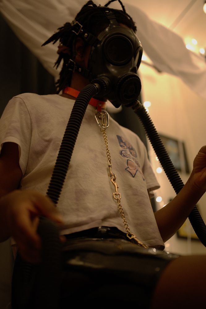 Myself in a s10 gasmask with tubes attached, orange collar with silver caribeaner and gold chain, a pastel sailor moon croptop shirt, and a latex skirt. I'm holding on of the tubes in my hands and am expressionless due to the blackout lenses on the gasmask. I'm in front of a softly lit background. 