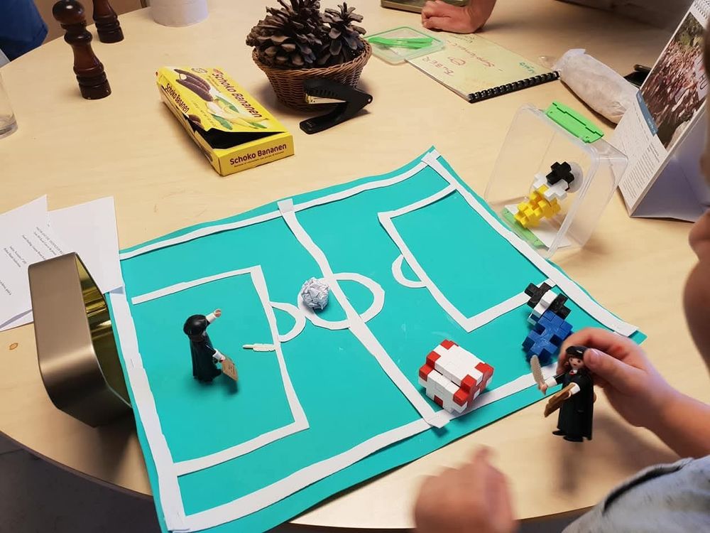 A soccer field made of paper, with the goals made of an empty candy tin and a tupperware. The ball is a crumpled piece of paper. The "players" are several Playmobil Martin Luthers and some blocks made of Plus Plus toys.
