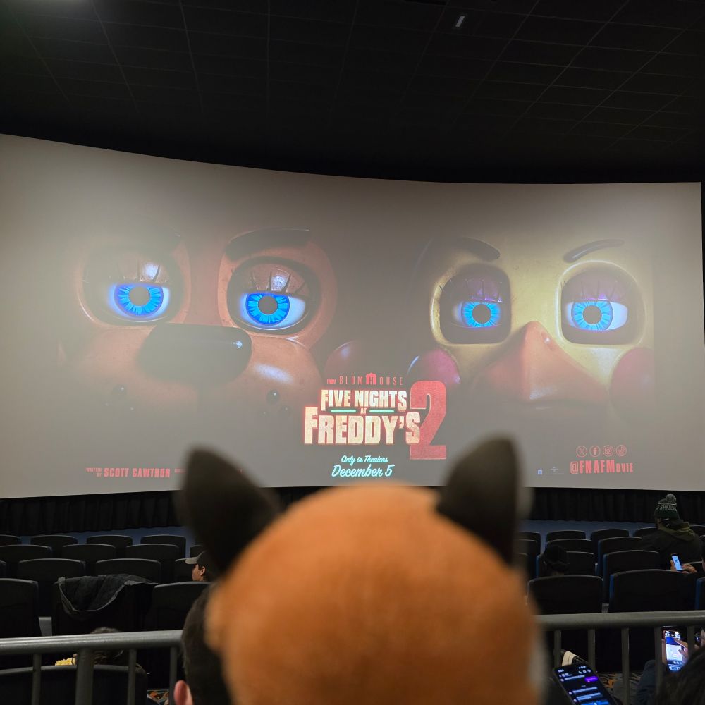 Raposa, a small stuffed fox, sits in front of a movie theater splash screen at a preview screening of #FNAFMovie only in theaters December 5, 2025