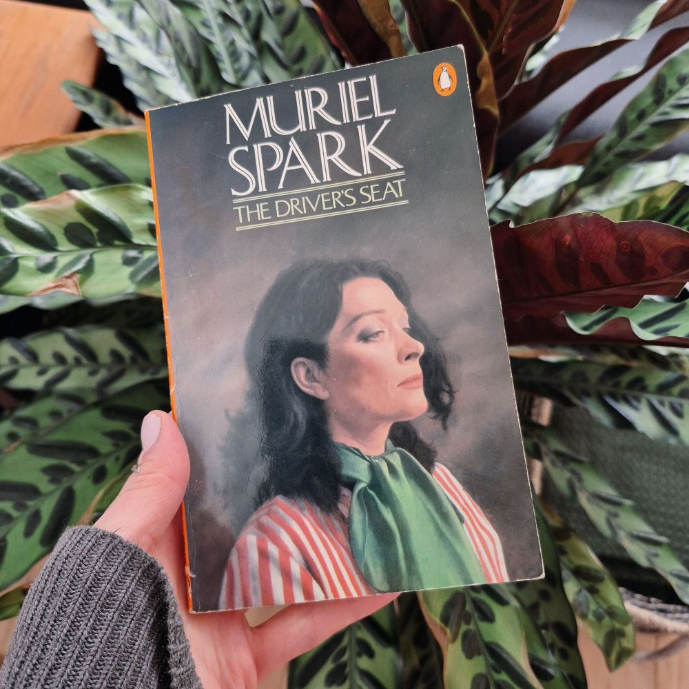 A copy of The Driver's Seat by Muriel Spark held in front of a plant with variegated leaves by a white person with a grey sleeve.