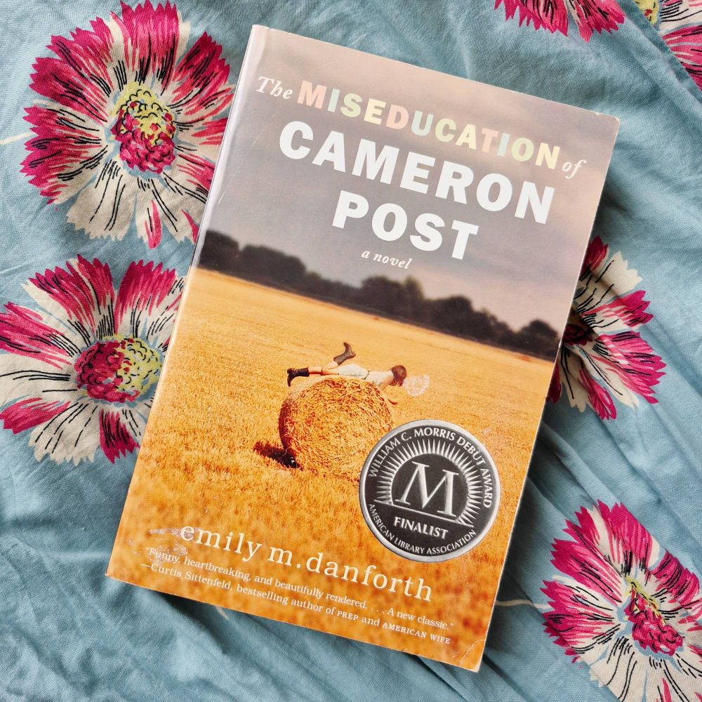 A copy of The Miseducation of Cameron Post on blue fabric with white and pink flowers. The book cover has a girl lying on a hay bale.