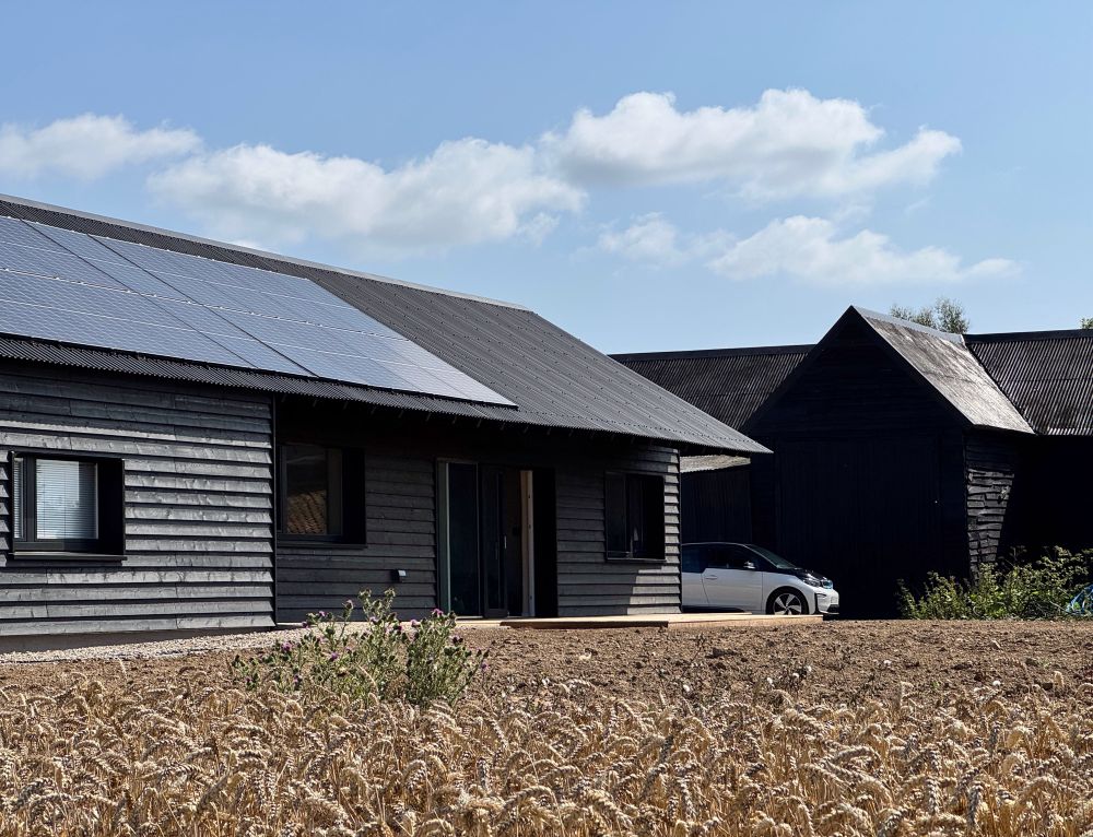Healthy and energy efficient single storey new home in Essex built with largely biobased materials such as timber, straw and clay. The building is finished with black materials including timber cladding and corrugated metal roof, paying homage to the existing farm buildings in the background.