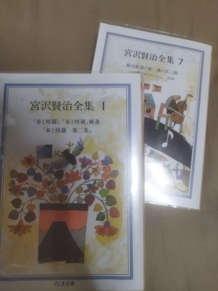 『宮沢賢治全集』（ちくま文庫）の1、7巻。前者の表紙絵は夕焼けの強い光を背にした富士山があしらわれた寸胴の花瓶に生けた花々と、それが織りなす紋様のなかに翼を広げた鳥の姿も。後者はセロ弾く人物（セロ弾きのゴーシュと思われる）と、道を挟んでそれを聴く様々な動物たち
