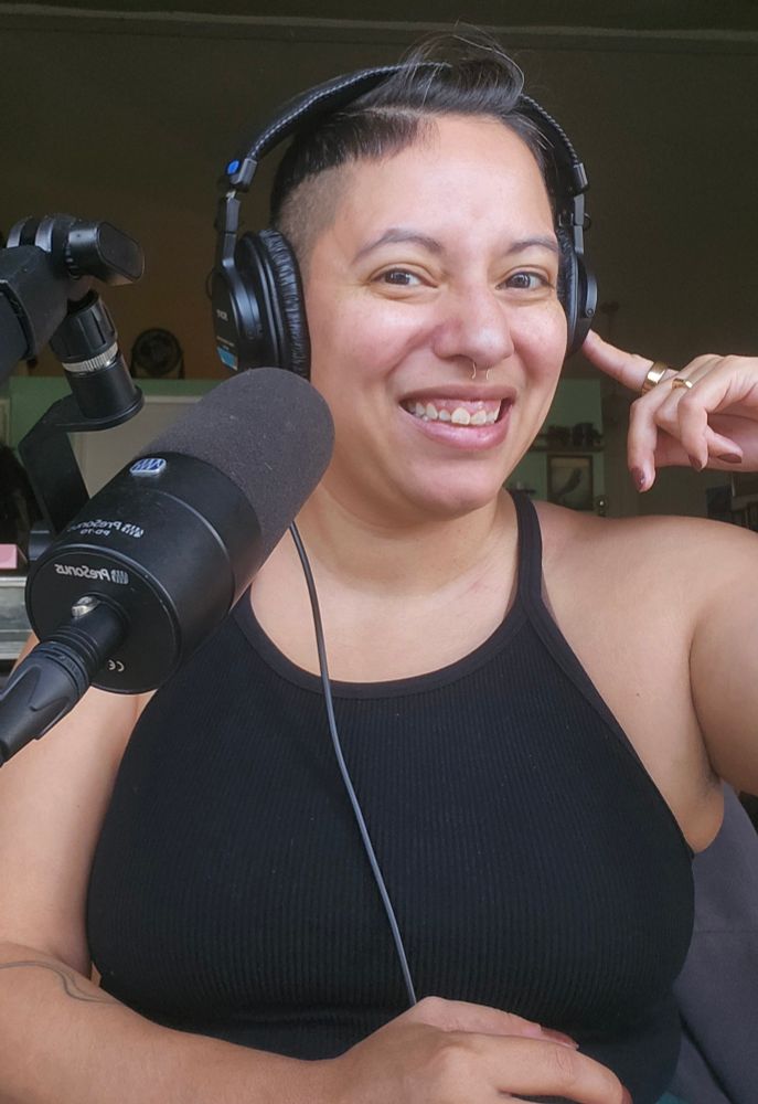 A woman with in a black top, with olive skin and a dark braided mohawk sits in front of a microphone, wearing headphones. She is smiling at the camera with her right index finger resting on the ear over her headphones. She has tattoos on her arms, rings on her fingers and a septum piercing. 
