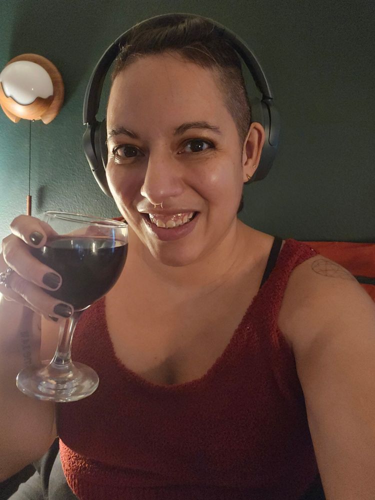 A woman with the sides of her head shaved holds a small glass of red wine. She has on a red top and a pair of green JLab Headphones. 