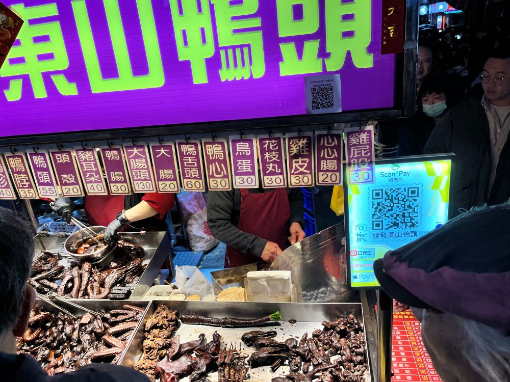 Booth with purple sign & Chinese green words, plus Chinese menu hanging over metal trays of darkly marinated and stewed or roasted meats