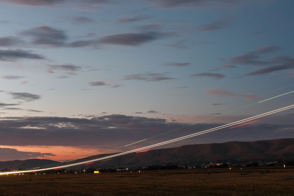 Early morning takeoff. Light trail from an Embraer with the sun just starting to turn the sky pink behind the ridge. Clouds scattered about the sky. Solid white light trails with dashed green and red from the beacons. 