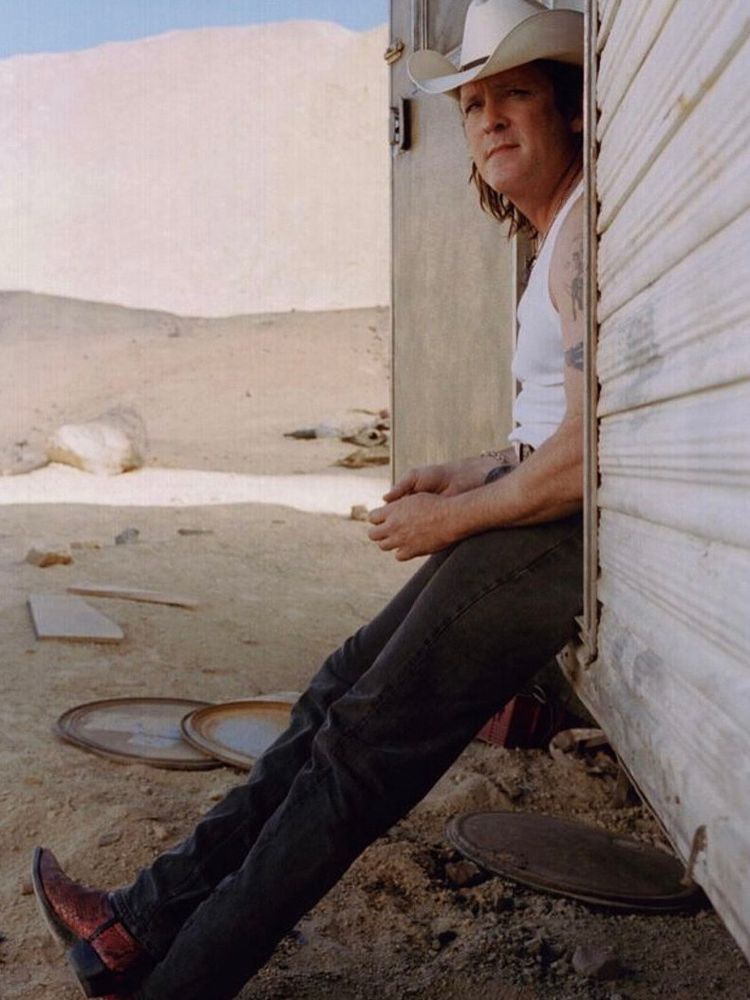Michael Madsen wearing cowboy boots, jeans, a white A shirt, and white cowboy hat. He is leaning against the doorway of a mobile home on the set of Kill Bill Vol. 2