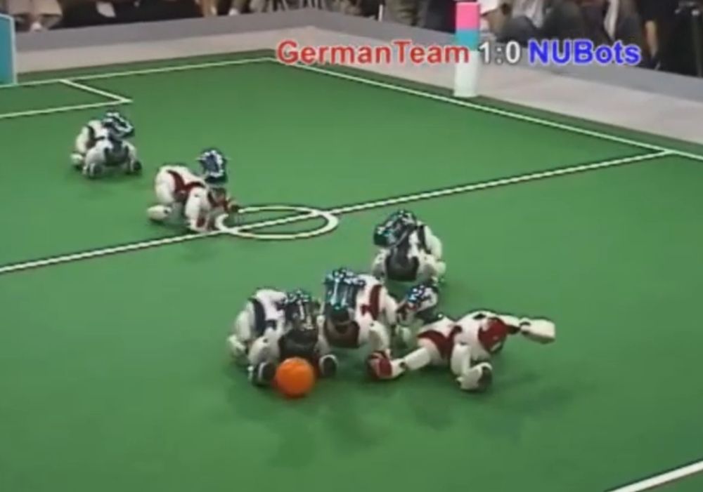 Robot soccer dogs playing in 2005, showing a robot diving to attempt to block a shot, while another robot manuevers around it