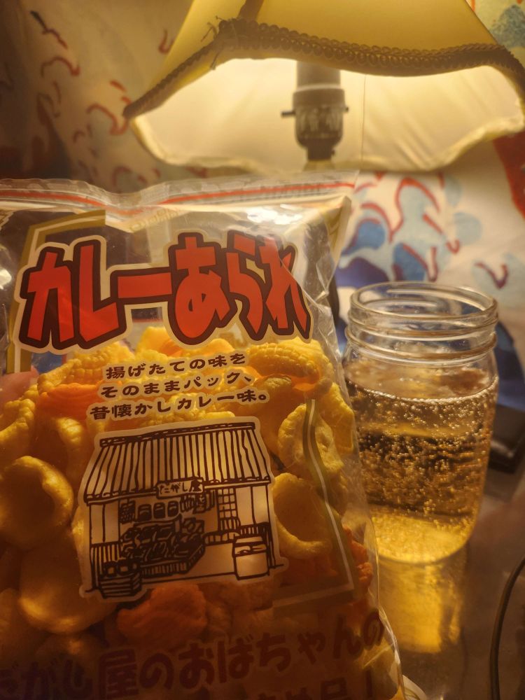 A picture of a bag of curry arare in front of a cup of ginger ale in front of a lamp with my wallet next to it. You can see a USB-C cable partially poke out from where my computer sits.