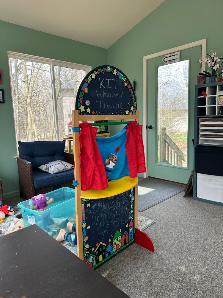 A room in a house with a tall windows, with a view of a railing, many bare trees, and another house not too far off. At center, a children's puppet theater with a chalkboard front, announcing Kit's Whimsical Theater. A blue sike (cyclorama) is framed by red curtains. A medieval knight puppet is at center. Visible behind the theater are a child's legs. The rest of the room is American middle class with IKEA shelving and table. Behind the theater is a plastic tote and a scattering of puppets, and a low rattan-like chair in front of one window.