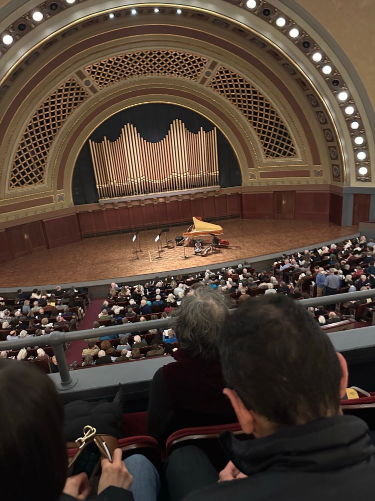 View from the mezzanine of a 3,500-seat concert hall (balcony not pictured). A seated audience waits before an oval stage holding a harpsichord, a bass, two cellos and a number of music stands. The back wall of the stage is an enormous pipe organ. (Hill Auditorium, University of Michigan)